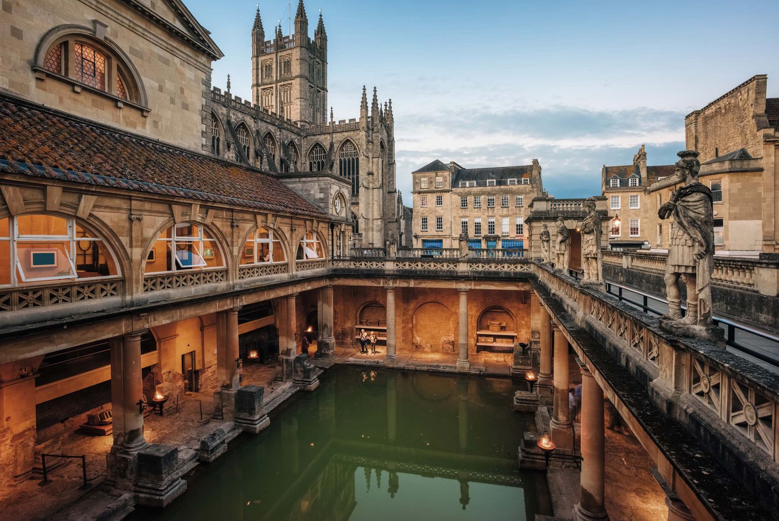 Historic Roman baths with green water, surrounded by ancient stone architecture and statues, with a large cathedral in the background at dusk.