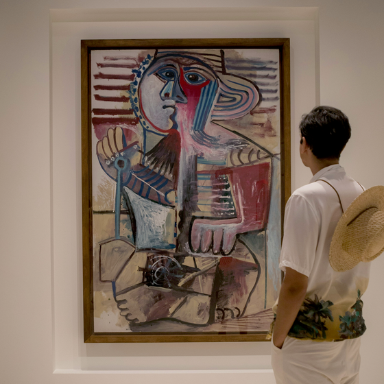 A person wearing a straw hat looks at a colorful abstract painting in a gallery.
