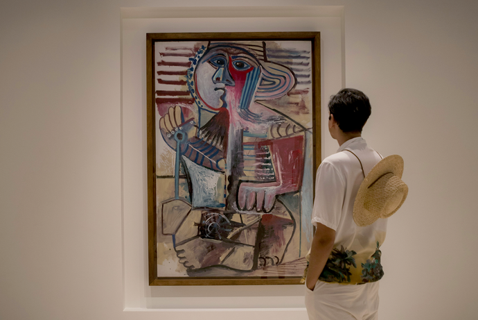 A person wearing a straw hat looks at a colorful abstract painting in a gallery.