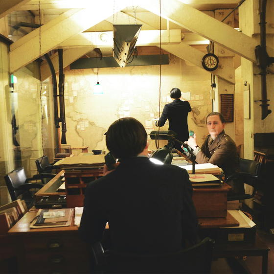 Historical office scene with three people at wooden desks, maps on the wall, and vintage lighting. One stands, writing on the map.