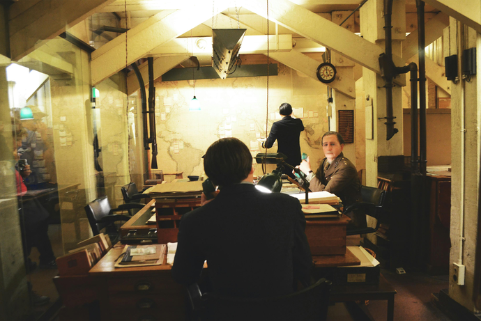 Historical office scene with three people at wooden desks, maps on the wall, and vintage lighting. One stands, writing on the map.
