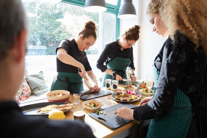 Four people in a kitchen, wearing aprons, preparing and garnishing dishes with a variety of colorful ingredients on a wooden table.