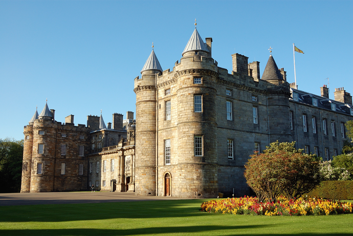 Historic stone castle with round towers, many windows, and a manicured garden with colorful flowers under a clear blue sky.