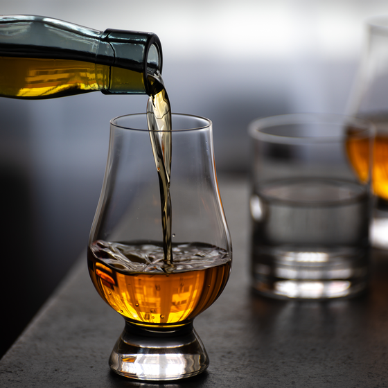 Whiskey being poured into a tulip glass on a table, with two more glasses nearby, one filled and the other empty.