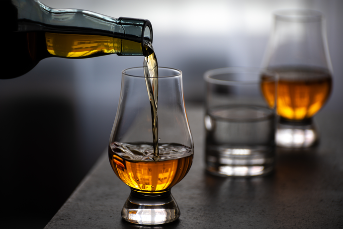 Whiskey being poured into a tulip glass on a table, with two more glasses nearby, one filled and the other empty.