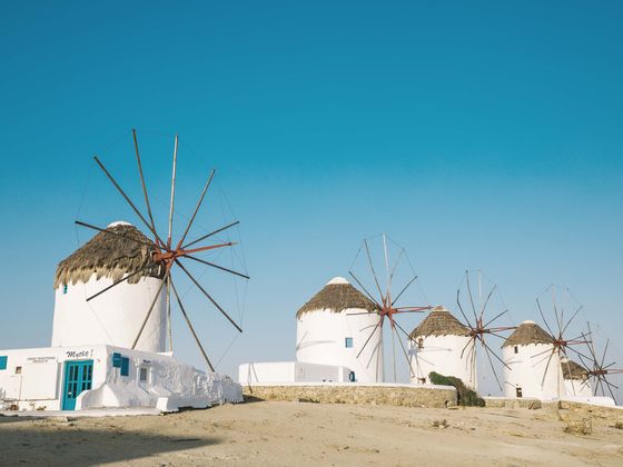 White windmills with thatched roofs against a clear blue sky, standing on a sandy hillside.