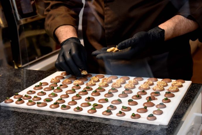 A person wearing black gloves arranges nuts on a tray of chocolate drops topped with pistachios, prepared on a black countertop.