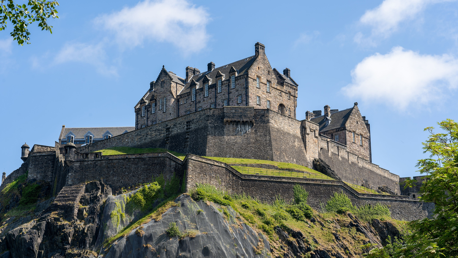 Stone castle atop a rocky hill against a blue sky, with green foliage in the foreground.