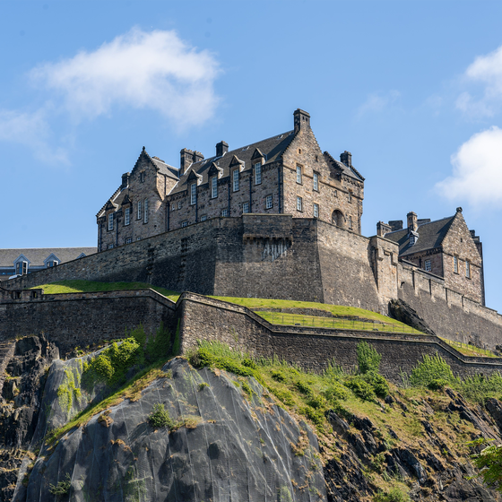 Stone castle atop a rocky hill against a blue sky, with green foliage in the foreground.