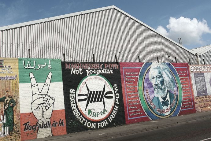 Murals on a corrugated metal wall featuring peace and historical themes, with barbed wire on top and a cloudy sky above.