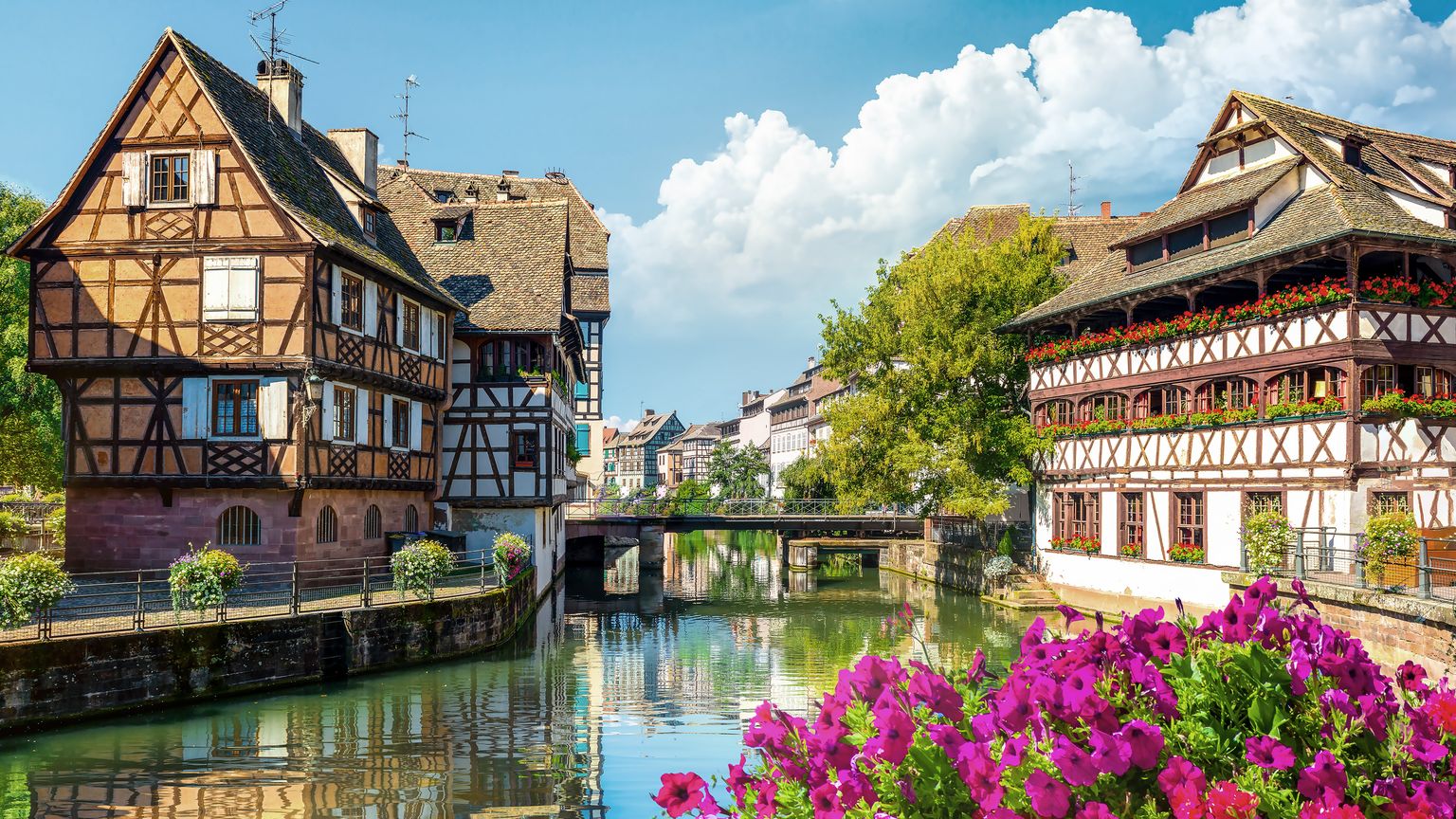 Canal water flows between historic half-timbered houses with tiled roofs, under a small bridge in the distance. Vibrant pink flowers bloom in the foreground, with more flower boxes visible on the balconies of the building on the right.