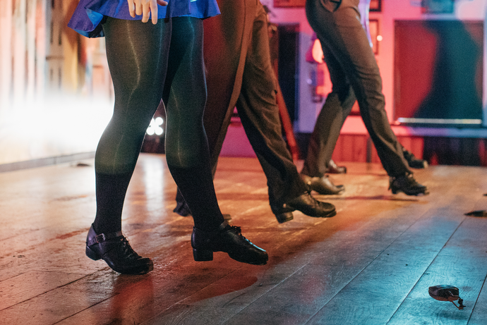 Dancers wearing tap shoes perform on a wooden stage, with focus on their feet in mid-step.