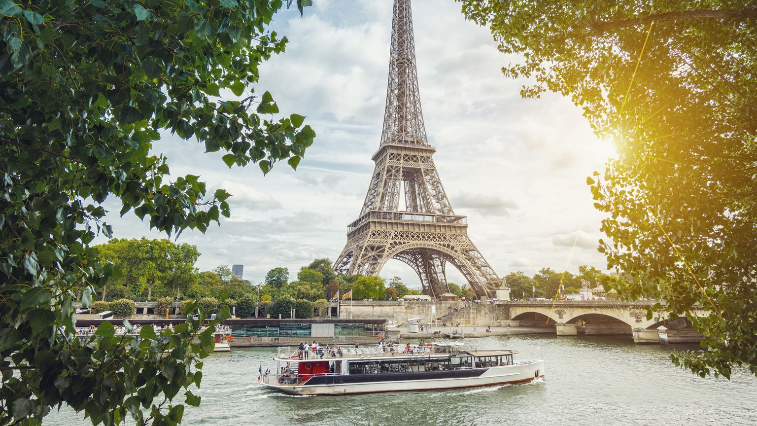 The Eiffel Tower stands against a cloudy sky, framed by overhanging tree branches in the foreground. A tour boat carrying passengers travels along the Seine River in front of a stone bridge.