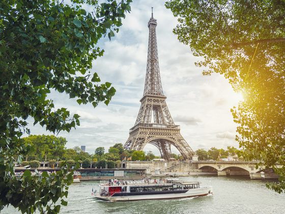 The Eiffel Tower stands against a cloudy sky, framed by overhanging tree branches in the foreground. A tour boat carrying passengers travels along the Seine River in front of a stone bridge.