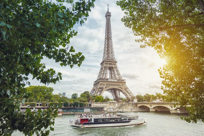 The Eiffel Tower stands against a cloudy sky, framed by overhanging tree branches in the foreground. A tour boat carrying passengers travels along the Seine River in front of a stone bridge.