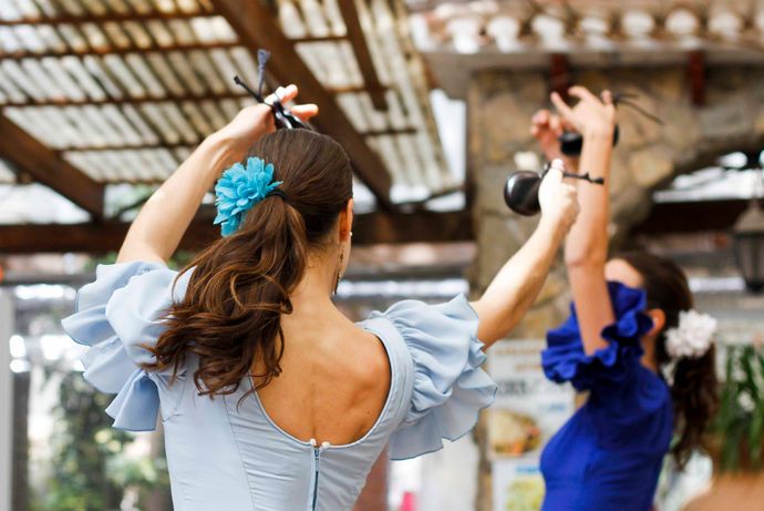 Two women in blue dresses dance flamenco with raised arms and castanets, under a pergola. One wears a blue hair flower, the other a white one.