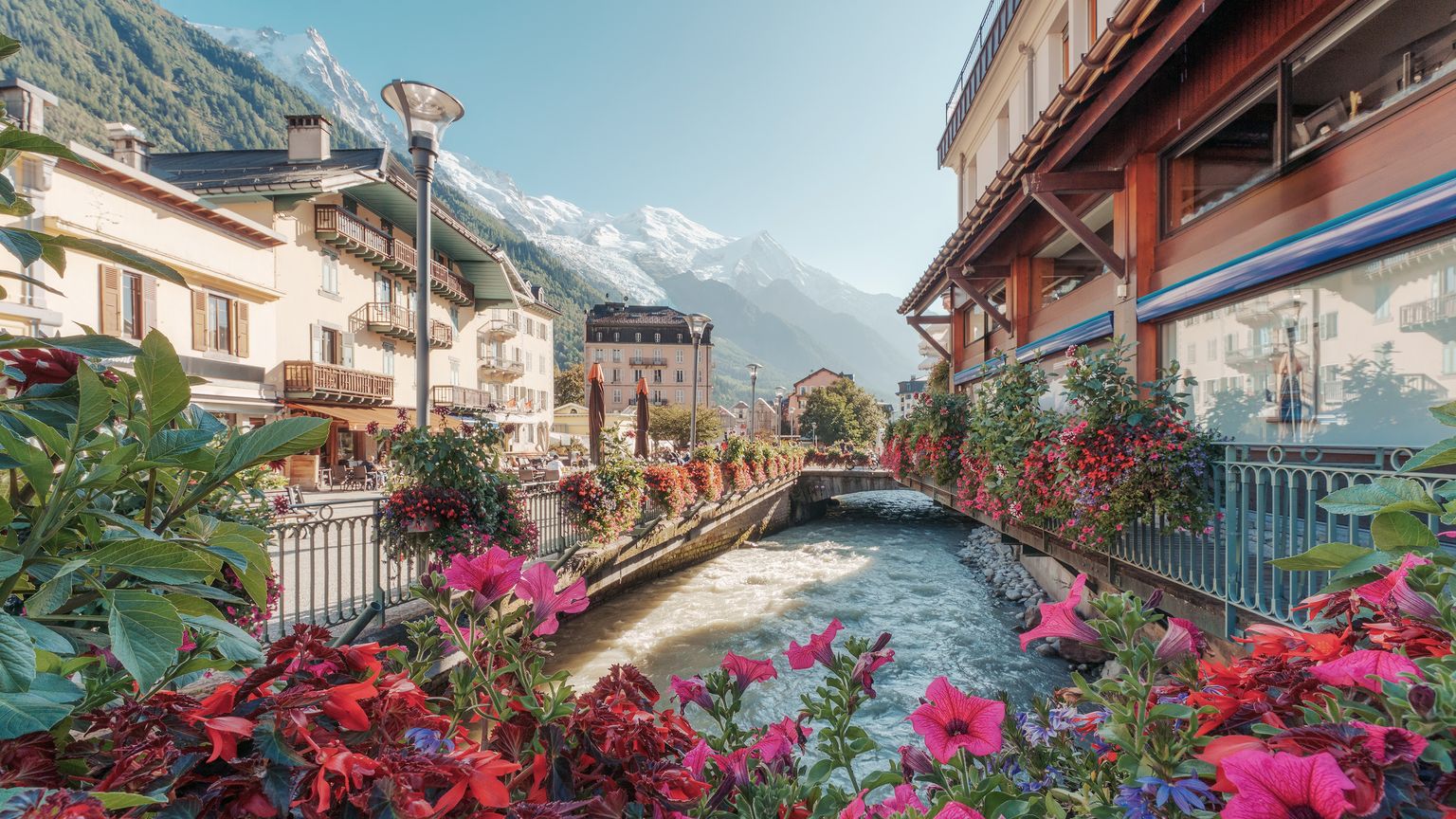 A river flows between buildings adorned with flower boxes in a town setting, with snow-capped mountains in the background. Pink and red flowers in the foreground frame the river view.
