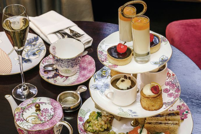 A tiered stand with assorted desserts and sandwiches, accompanied by tea cups and a champagne glass on a table set for afternoon tea.