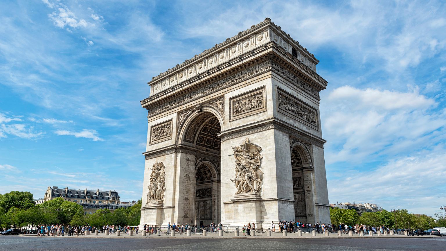The Arc de Triomphe in Paris on a clear day, surrounded by people and trees, under a blue sky with scattered clouds.