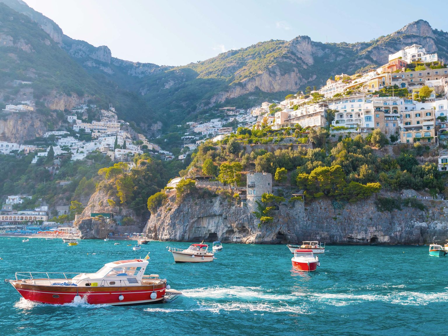 Colorful boats on turquoise water with cliffside homes in Positano, Italy, under a clear blue sky.