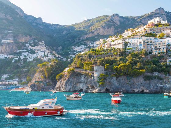 Colorful boats on turquoise water with cliffside homes in Positano, Italy, under a clear blue sky.