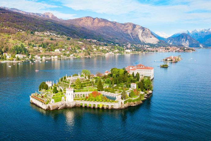 Scenic view of an island with lush gardens and historic buildings, surrounded by a serene lake and mountains under a blue sky.