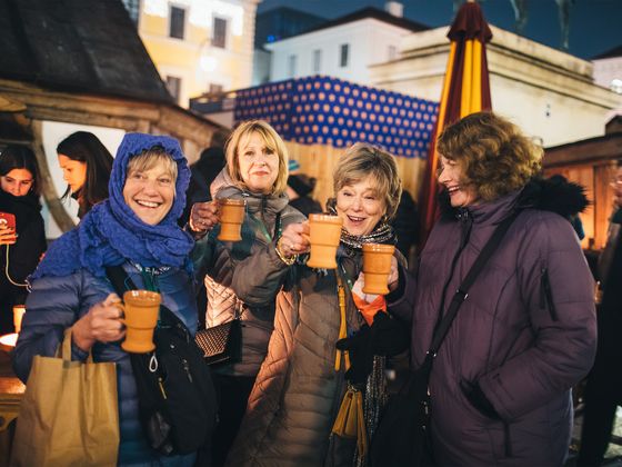 Four women in winter coats hold mugs and smile at an outdoor market at night, surrounded by festive lights and stalls.