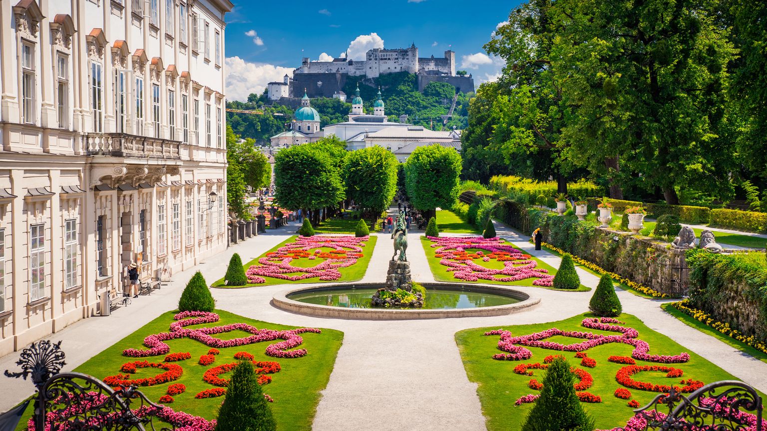 A vibrant garden with manicured flower beds, a central fountain, and a grand building, with a hilltop fortress in the background under a blue sky.