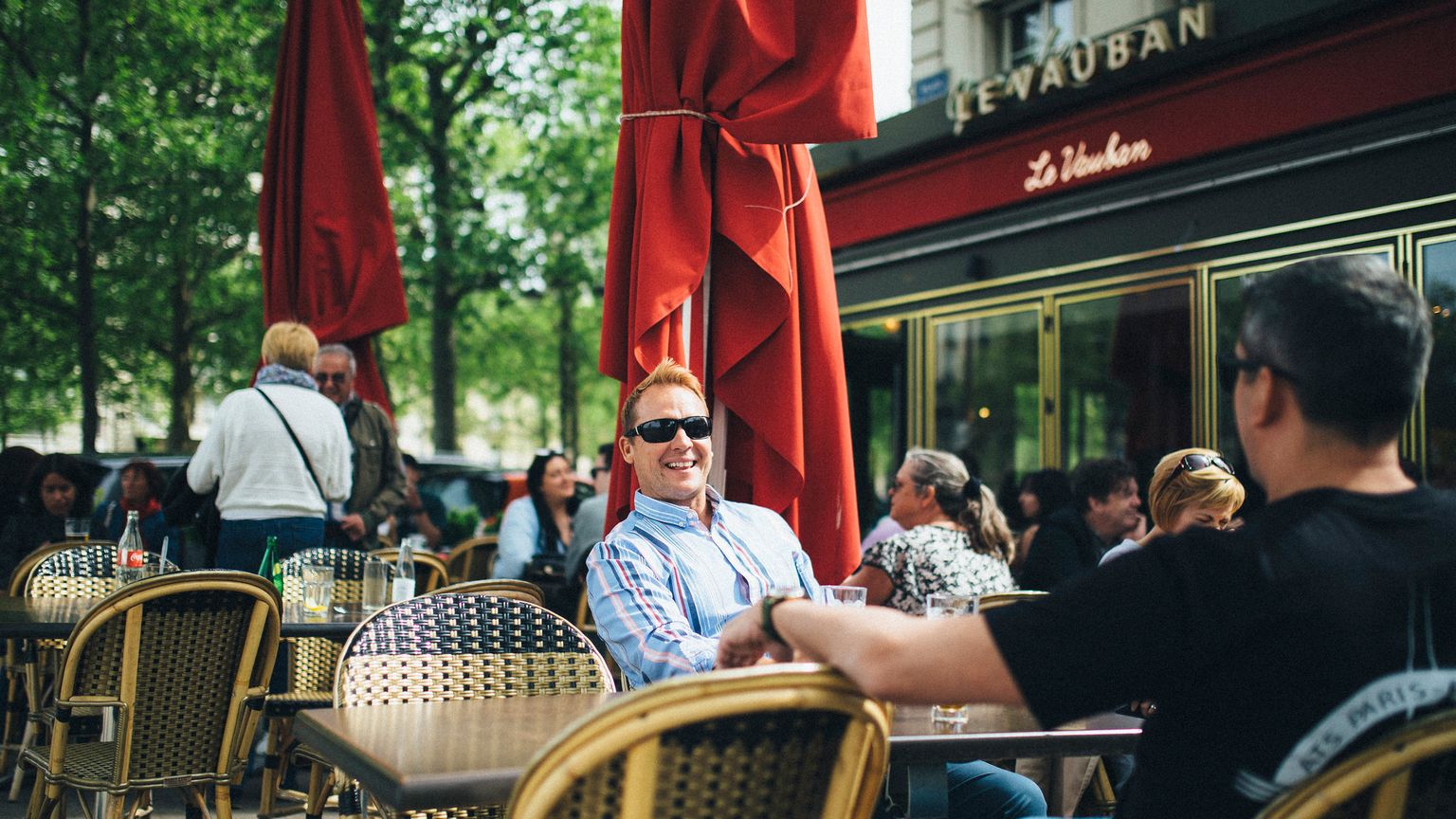 People sitting at an outdoor café with wicker chairs and red umbrellas, enjoying a sunny day. The scene is lively and relaxed.