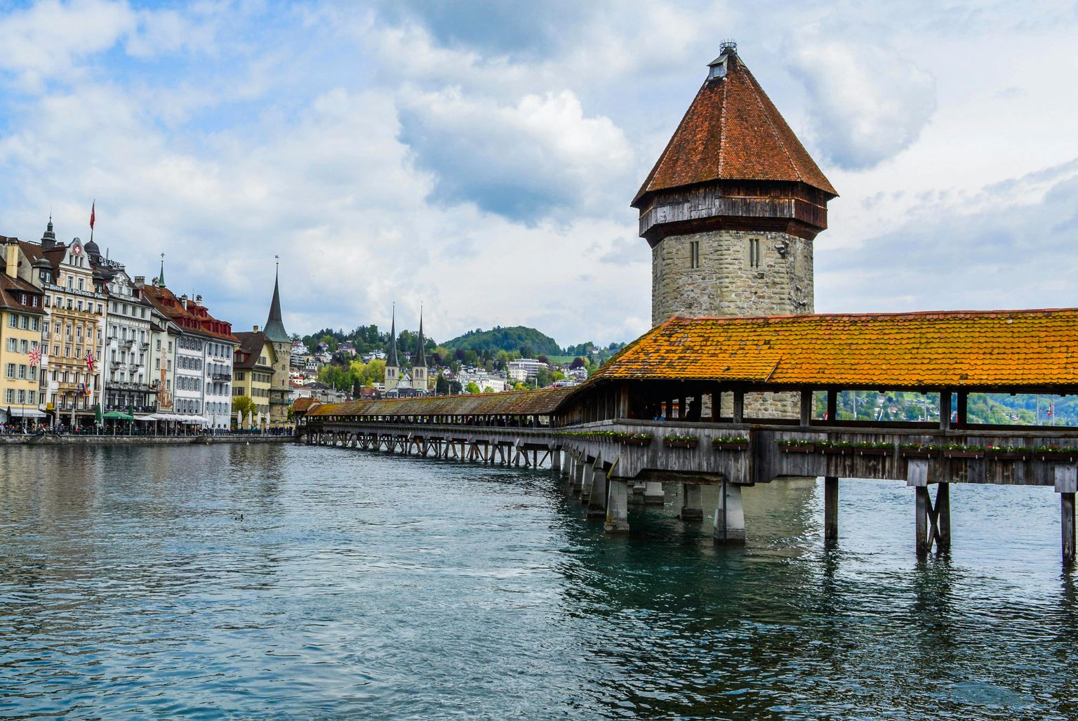 Historic wooden Chapel Bridge spans a river with a stone tower, surrounded by colorful buildings and distant hills under a partly cloudy sky.