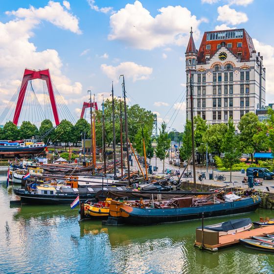 Historic boats docked at a scenic harbor with a red bridge and an ornate building in the background, surrounded by lush greenery on a sunny day.