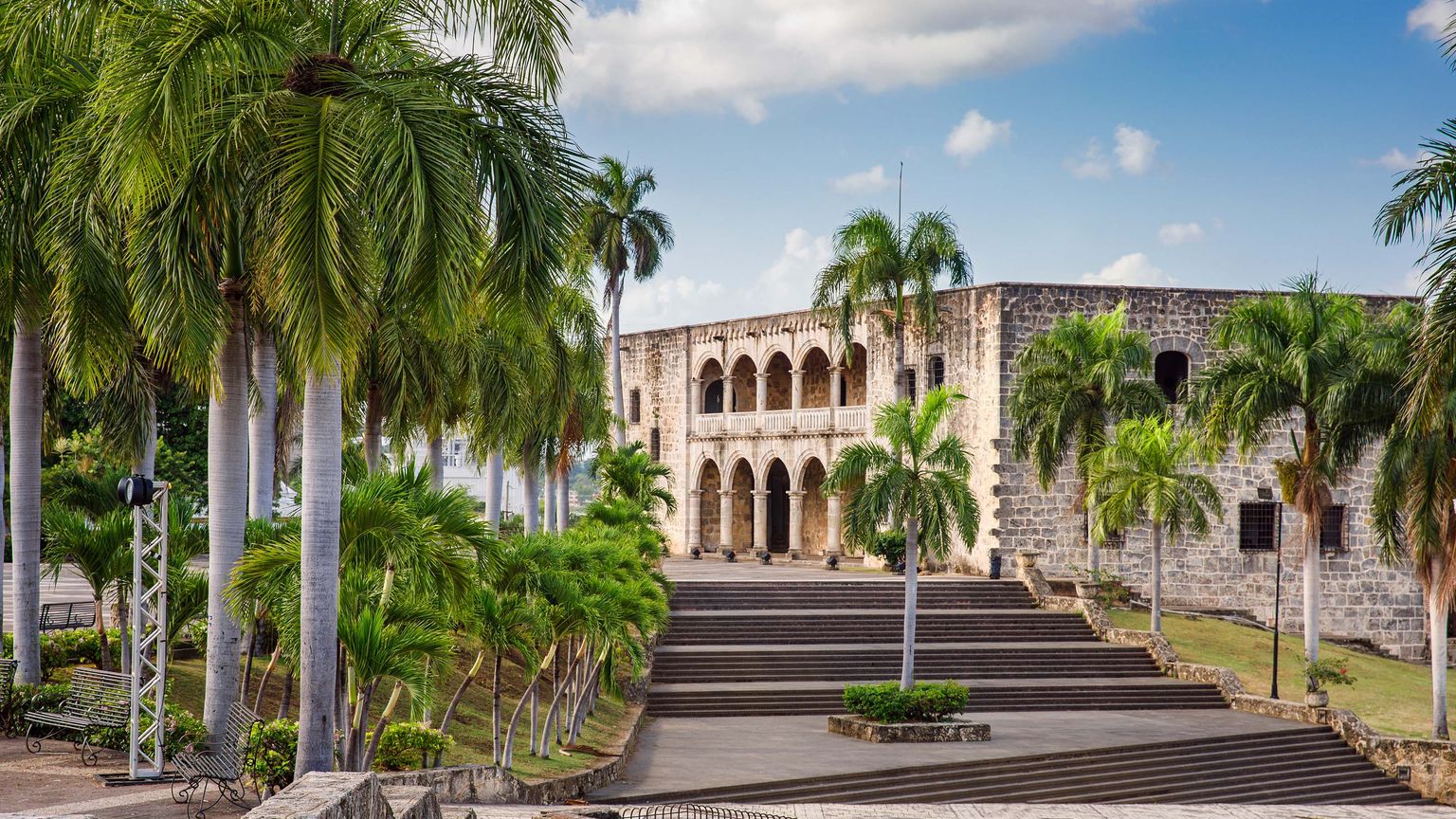 Historic stone building with arched balconies, surrounded by palm trees and steps, under a blue sky with clouds.