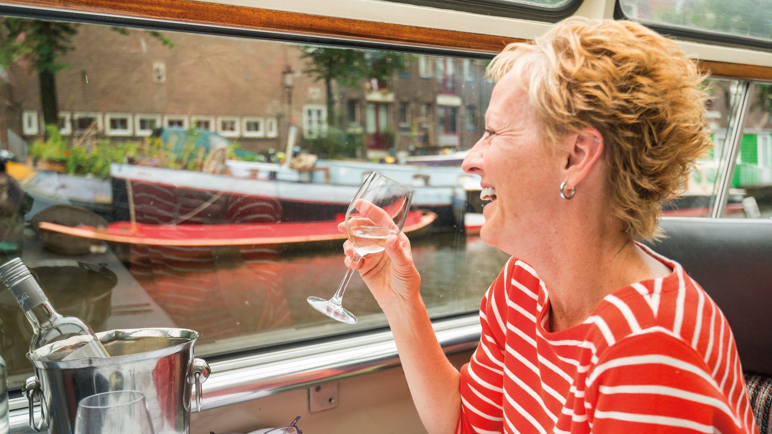 A smiling woman in a red striped shirt enjoys a glass of wine on a boat, with a canal and boats visible through the window.