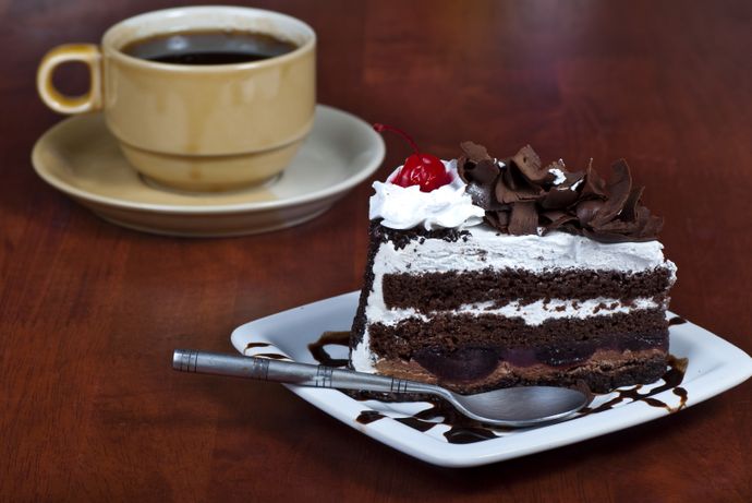 A slice of chocolate cake with whipped cream, a cherry, and chocolate shavings on a white plate with a spoon, next to a cup of coffee.