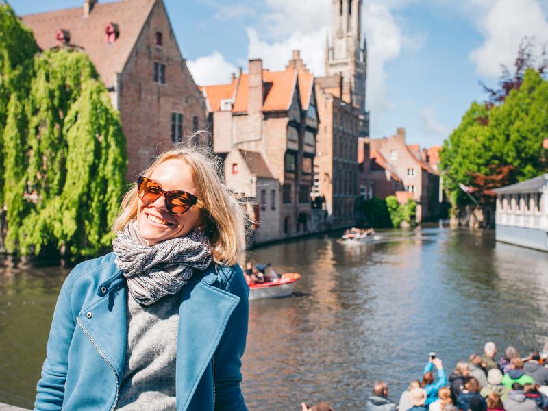 Smiling woman in sunglasses stands by a scenic canal in Bruges, with historic buildings and trees in the background on a sunny day.