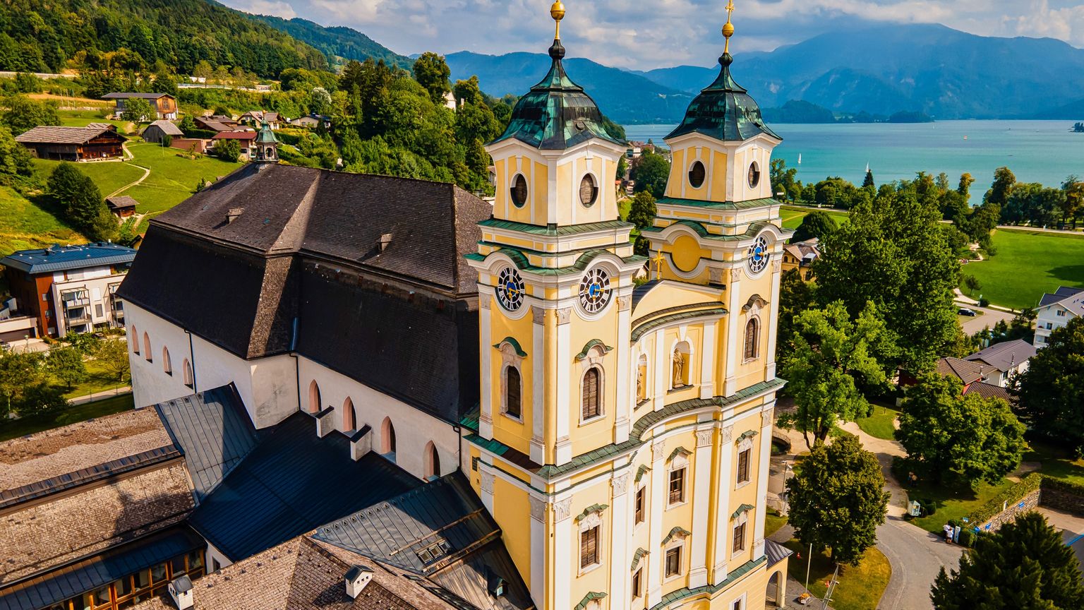 Aerial view of a yellow baroque church with twin towers, surrounded by lush greenery and overlooking a turquoise lake and distant mountains.
