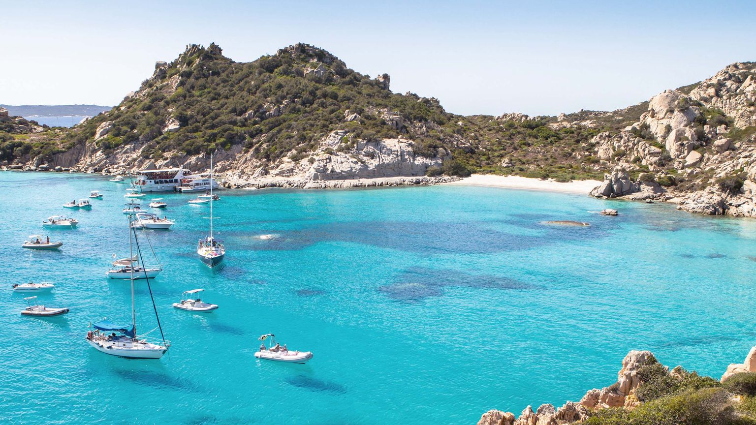 Turquoise bay with multiple sailboats and motorboats, surrounded by rocky hills and lush greenery under a clear blue sky.