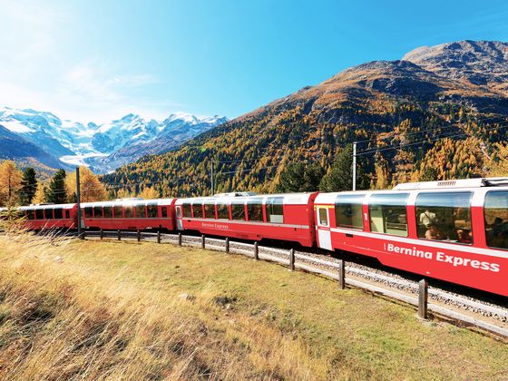 Red Bernina Express train travels through scenic autumnal mountain landscape with snow-capped peaks and colorful foliage.