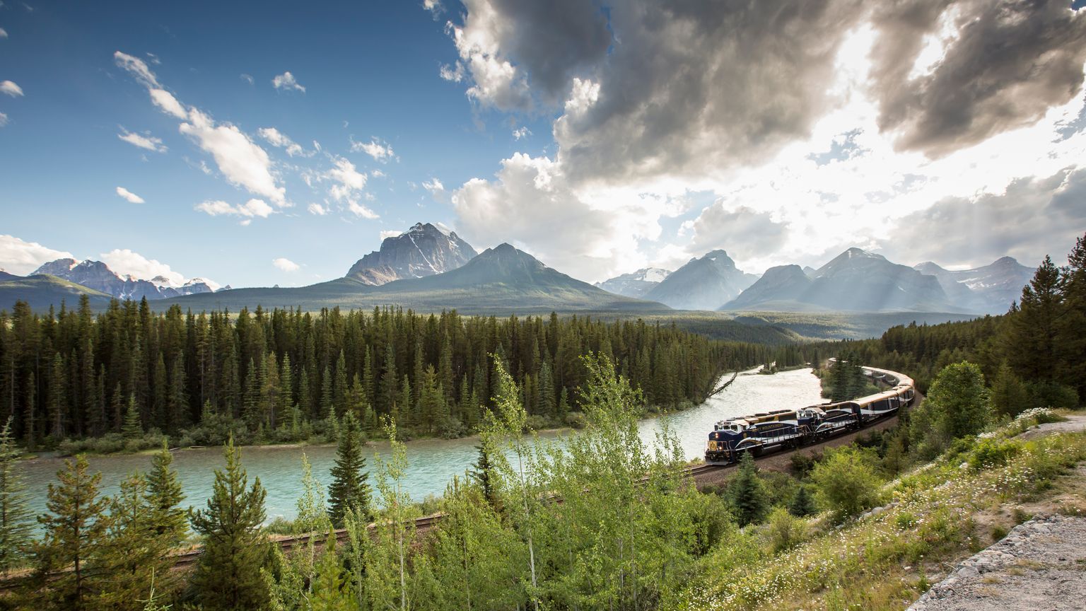 A train travels along a winding track by a river, surrounded by lush forest and majestic mountains under a partly cloudy sky.