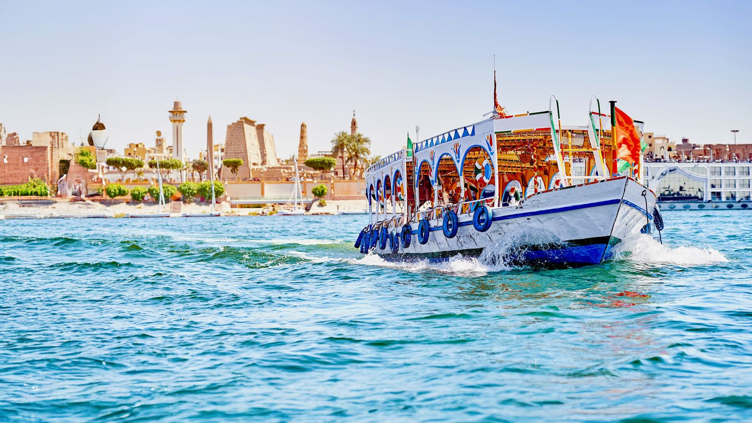 A vibrant boat sails on a river with an ancient temple complex in the background under a clear blue sky.