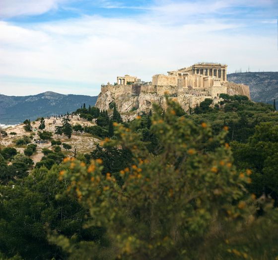 View of the Acropolis in Athens, Greece, with the Parthenon atop a rocky hill, surrounded by lush greenery and distant mountains under a blue sky.