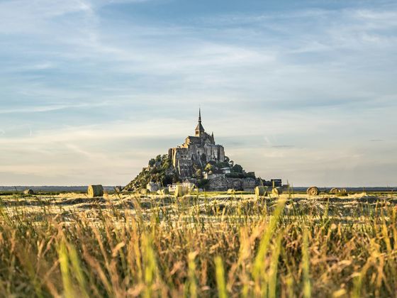 A historic abbey sits atop a rocky island surrounded by hayfields under a clear sky.