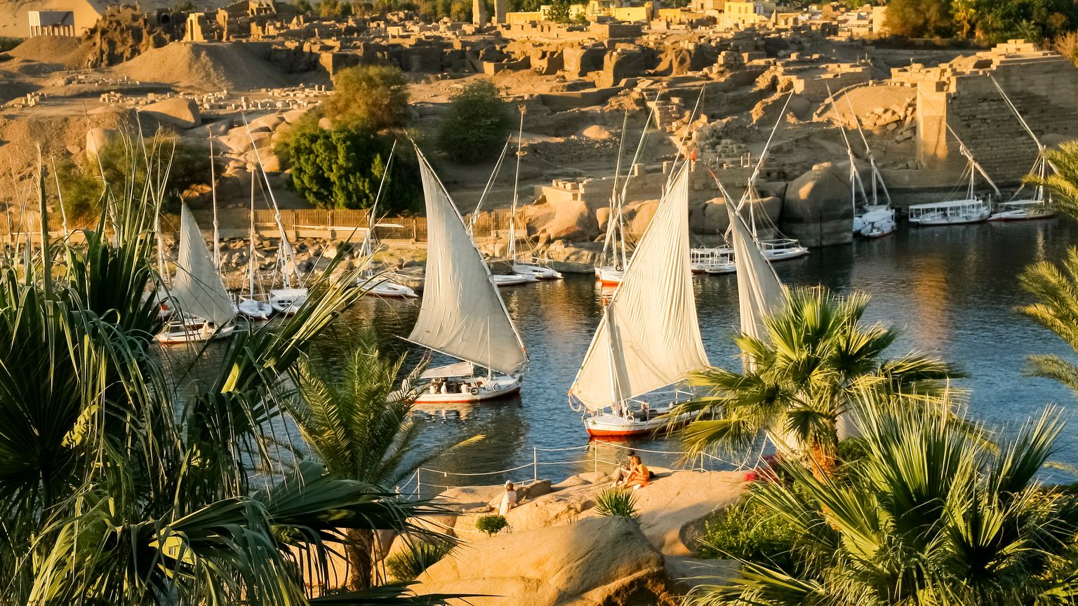 Sailboats with white sails on the Nile river bordered by palm trees, with ancient ruins and desert hills in the background under a clear sky.