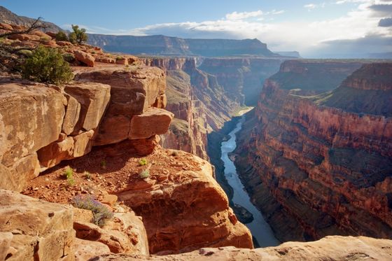 Majestic view of the Grand Canyon with rocky cliffs and a winding river below, under a vast blue sky with scattered clouds.