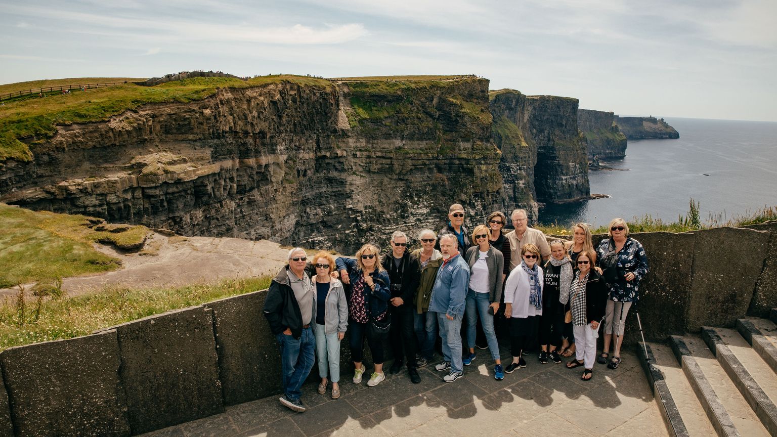A group of people posing in front of the Cliffs of Moher, Ireland, with the ocean and cliffs in the background under a partly cloudy sky.