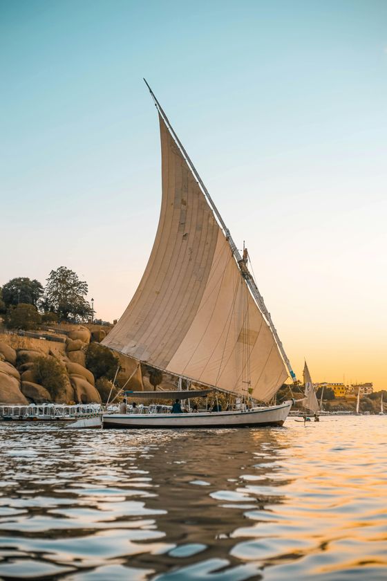 A traditional sailboat with large sails glides on calm water at sunset, with rocky banks and a clear sky in the background.