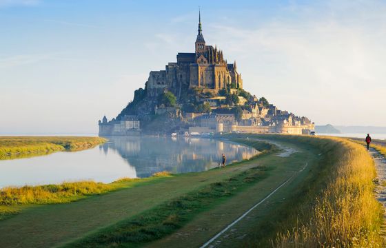 A distant view of Mont Saint-Michel, a historic island commune in Normandy, France, surrounded by water and a grassy path.