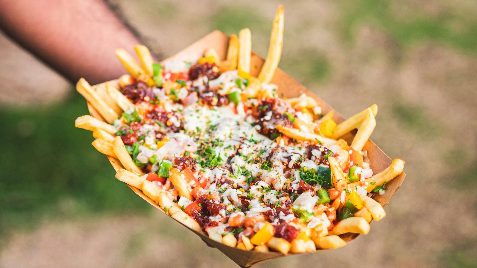 A hand holding a cardboard tray filled with french fries topped with melted cheese, chopped tomatoes, green peppers, onions, and herbs.