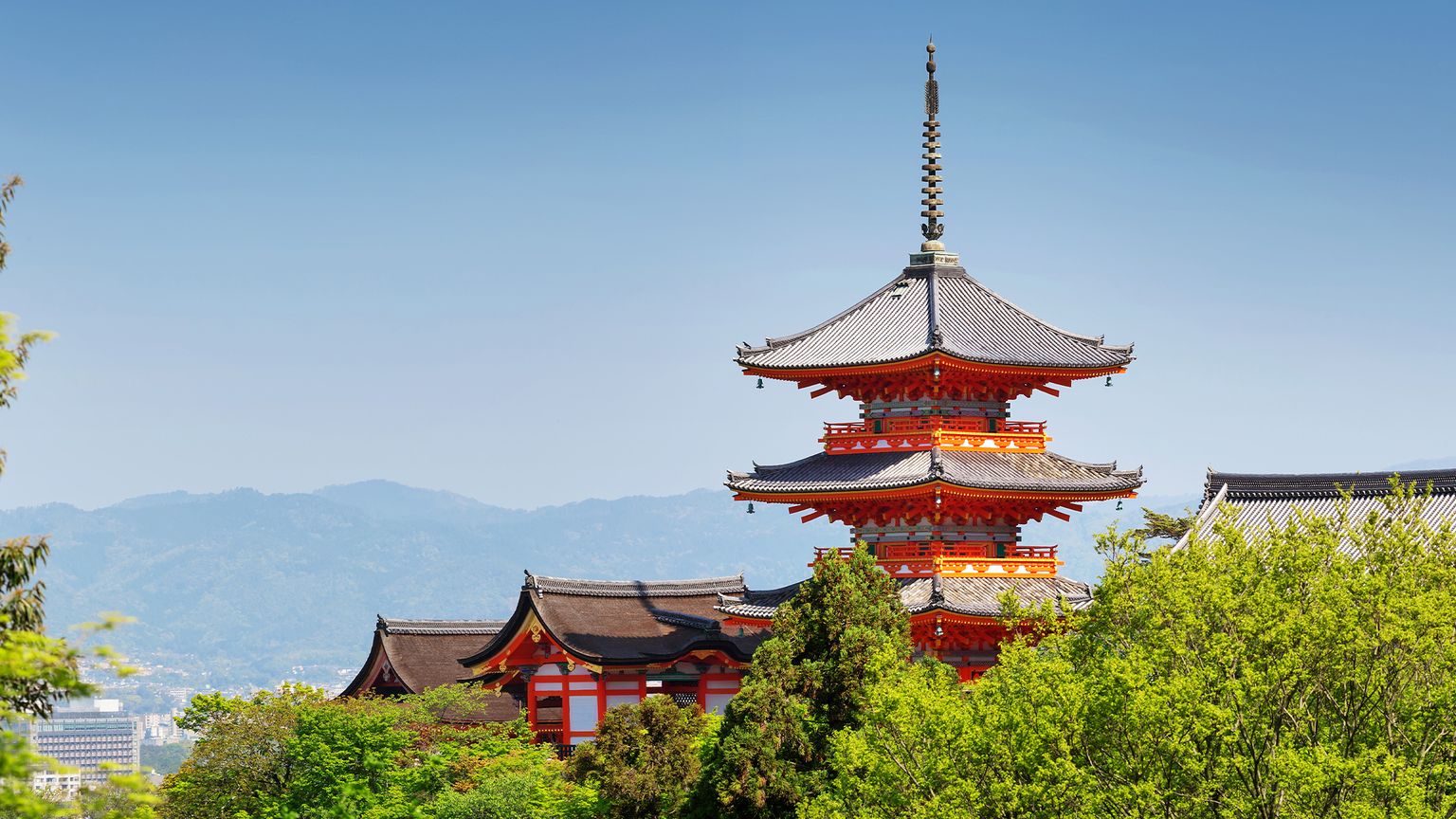 A scenic view of Kiyomizu-dera temple with a red pagoda surrounded by lush greenery and distant mountains under a clear blue sky.