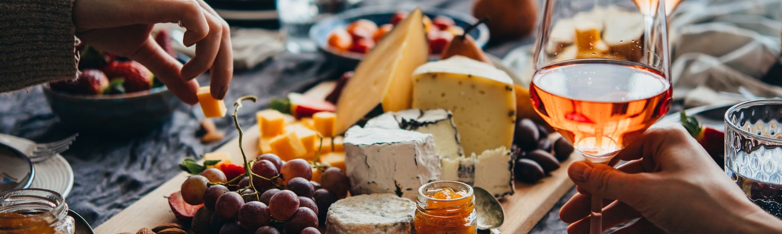 A table with assorted cheeses, grapes, berries, olives, and a glass of rosé wine; hands reaching for food.